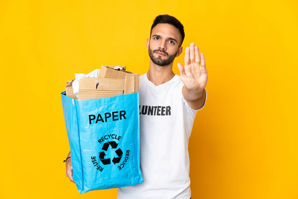 Joven hombre caucásico sosteniendo una bolsa de reciclaje llena de papel para reciclar aislado sobre fondo blanco haciendo gesto de parada - Foto, imagen