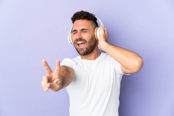 Caucasian man isolated on purple background listening music and singing - Foto, imagen
