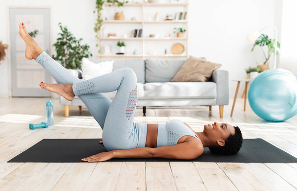 Entrenamiento deportivo en casa. Atractiva jovencita tumbada sobre esterilla de yoga y cicing en el aire, haciendo ejercicios abdominales en salón - Foto, imagen
