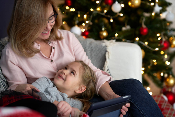 Grandmother with granddaughter using tablet while relaxing on sofa                                - Photo, Image