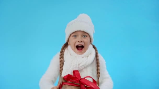 счастливая девушка в трикотажном белом зимнем наряде с подарочной коробкой изолированы на синий - Кадры, видео