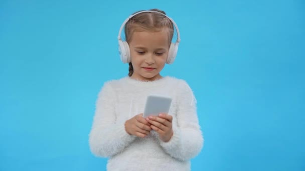 Mädchen im flauschigen Pullover und Kopfhörer mit Smartphone isoliert auf blau - Filmmaterial, Video