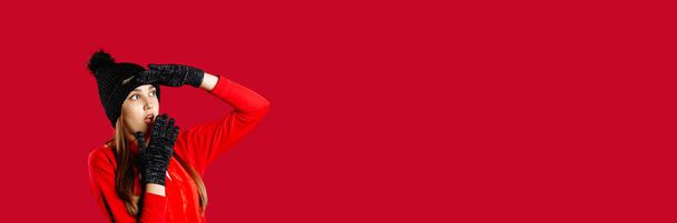 Banner, long format. Surprised girl holding a hand to the face in a winter hat and mittens on an isolated red background looks to the side. High quality photo - Fotó, kép