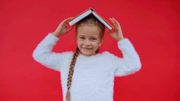 Ευτυχισμένο κορίτσι σε χνουδωτό πουλόβερ βάζοντας το βιβλίο στο κεφάλι απομονωμένο στο κόκκινο - Πλάνα, βίντεο