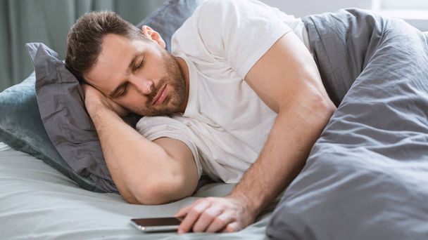 Людина, яка спить, тримає смартфон лежачи у ліжку в приміщенні (панорама) - Фото, зображення