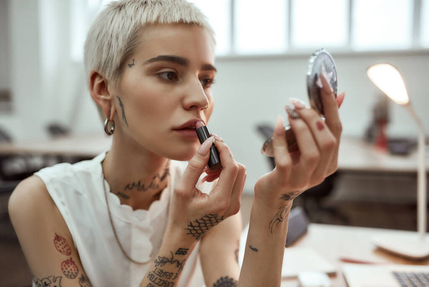 Doing make up. Portrait young and attractive blonde tattooed woman applying lipstick while sitting in the modern office - Foto, Imagen