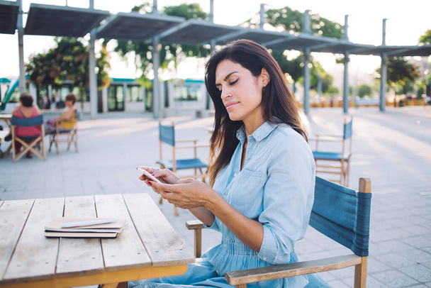 Serious brunette caucasian woman in casual wear resting on cafe terrace during weekend browsing social networks and reading news on mobile phone, pensive female millennial installing app on cellular - Фото, зображення