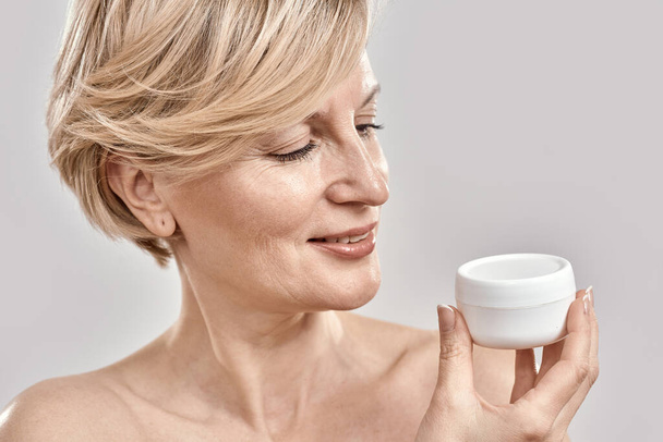 Close up portrait of beautiful middle aged woman holding, looking at moisturizing cream jar while posing isolated over grey background - Foto, Bild