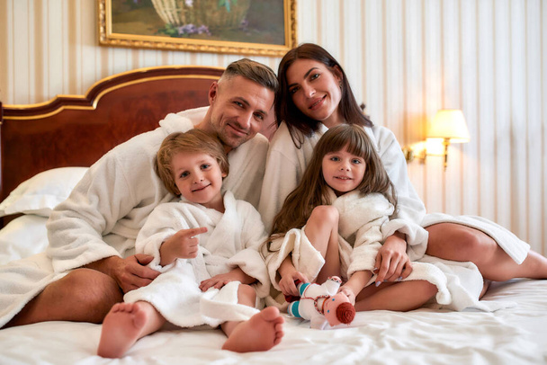Een levensstijl ervaring. Blanke ouders en twee kinderen in witte badjassen glimlachend naar de camera terwijl ze op het bed zitten in een luxe hotelkamer. Familie, reizen, resort, vakantie concept - Foto, afbeelding