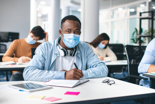 Schwarzer männlicher Student in Maske sitzt am Schreibtisch im Klassenzimmer - Foto, Bild