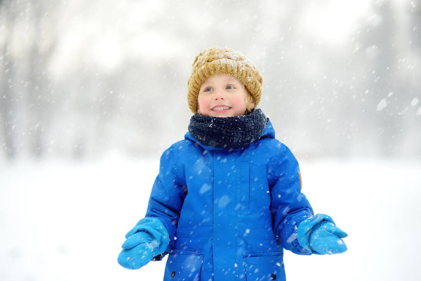 Little boy having fun playing with fresh snow during snowfall. Baby catching snowflakes on gloves. Kid dressed in warm clothes, hat, hand gloves and scarf. Active winter outdoors leisure for child - Foto, Bild
