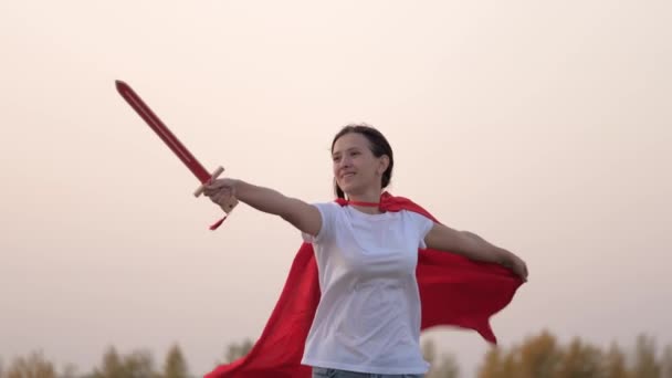 Dětští rytíři. Svobodná dívka v červeném plášti běhá s meči v ruce přes pole a hraje si na středověkého rytíře. Mladá dívka si hraje na superhrdiny. Děti se perou mečem. Šťastné dětství - Záběry, video