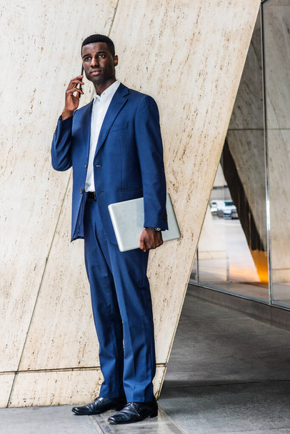 Молодой афро-американский бизнесмен в синем костюме, белой майке, кожаных туфлях, с ноутбуком в руках, стоит в колонке на улице возле офиса в Нью-Йорке, разговаривает по мобильному телефону снаружи - Фото, изображение