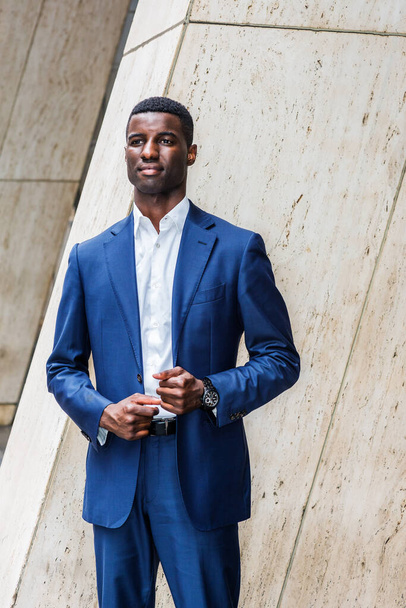 Genç Afrikalı yakışıklı iş adamı mavi takım elbiseli, beyaz atlet, kol saati, ofis binasının dışında köşesinde duruyor, kaşlarını çatmış, bekliyor, bekliyor. - Fotoğraf, Görsel