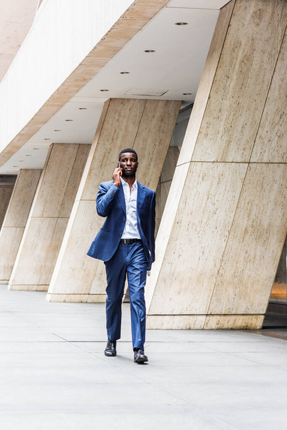 Empresário afro-americano viajando, trabalhando em Nova York, vestindo terno azul, camiseta branca, sapatos de couro, carregando computador portátil, andando na rua fora do prédio de escritórios, falando por telefone - Foto, Imagem