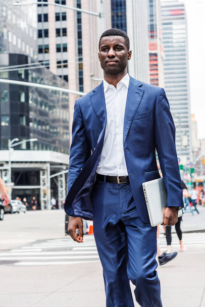 Jeune homme d'affaires afro-américain voyageant à New York, portant un costume bleu, un maillot blanc, portant un ordinateur portable, marchant dans une rue animée avec de hauts bâtiments dans le Midtown de Manhattan - Photo, image