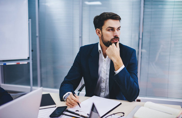 Contemplative male boss dressed in formal clothing sitting at table desk and thoughtful looking away while doing accounting job, pensive executive manager thinking about details of financial report - Фото, зображення