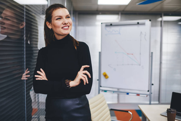 Confident cheerful female leader in stylish clothing looking away and smiling during working time in company, happy corporate director enjoying own occupation and business lifestyle in office - Foto, imagen