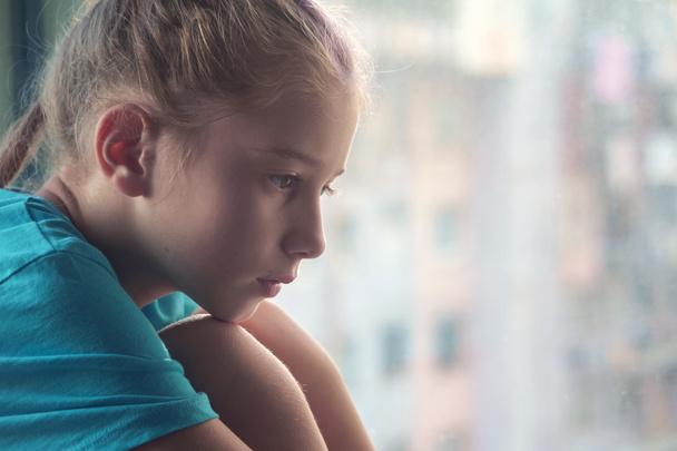 Uma menina triste de olhos castanhos sentada no peitoril da janela com fundo ao ar livre borrado - Foto, Imagem