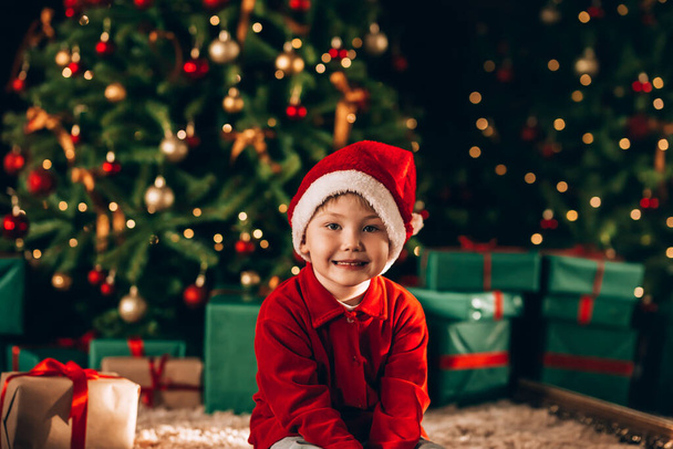 The boy has a santa hat on his head. In the background there is a large decorated Christmas tree with gifts in green packaging and a bow. Christmas mood. - Foto, afbeelding