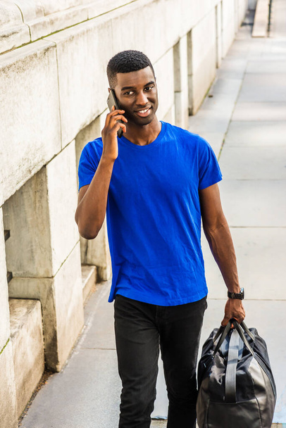 Terug naar school. Jonge Afro-Amerikaanse student studeert in New York, draagt blauw T-pak, zwarte broek, draagt leren tas, praat op mobiele telefoon, loopt buiten kantoorgebouw op de campus - Foto, afbeelding