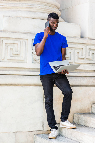Jeune étudiant afro-américain étudiant à New York, portant T-shirt bleu, pantalon noir, baskets, debout sur les escaliers contre le mur sur le campus, travaillant sur un ordinateur portable, parlant sur un téléphone portable - Photo, image