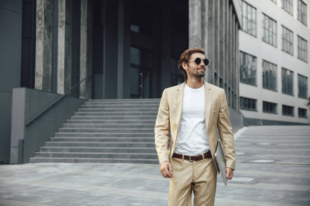 Vorderansicht eines erfolgreichen jungen Geschäftsmannes in trendigem Anzug und Sonnenbrille mit modernem Laptop, während er in der Nähe eines Bürogebäudes steht. Konzept von Errungenschaften und Technologie. - Foto, Bild