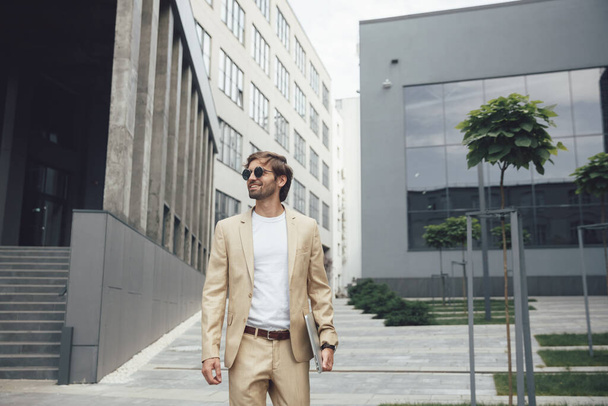 Portrait of smiling handsome man in stylish business suit and sunglasses standing near modern building with personal laptop in hands. Satisfied and successful person. - Zdjęcie, obraz