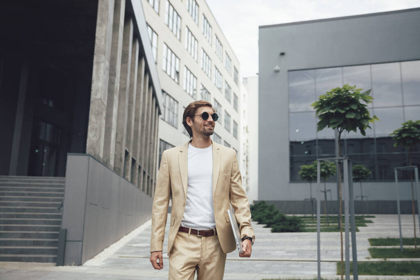 Μπροστά άποψη του ευτυχισμένος γενειοφόρος επιχειρηματίας περπάτημα με προσωπικό φορητό υπολογιστή στο δρόμο. Νεαρός με μαύρα γυαλιά ηλίου και μπεζ κοστούμι που χρησιμοποιεί για την εργασία του μοντέρνα συσκευή. - Φωτογραφία, εικόνα