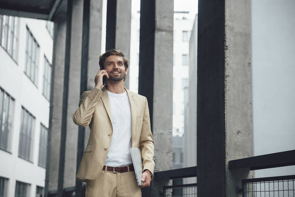 Впевнений бізнесмен в офіційному вбранні говорить на мобільному телефоні, стоячи на відкритому повітрі з ноутбуком в руці. Щасливий бородатий хлопець використовує сучасні гаджети для віддаленої роботи
. - Фото, зображення