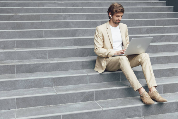 Przystojny brodaty mężczyzna w stylowym beżowym garniturze pracujący na bezprzewodowym laptopie siedząc na schodach. Pojęcie ludzi i nowoczesnej technologii. - Zdjęcie, obraz