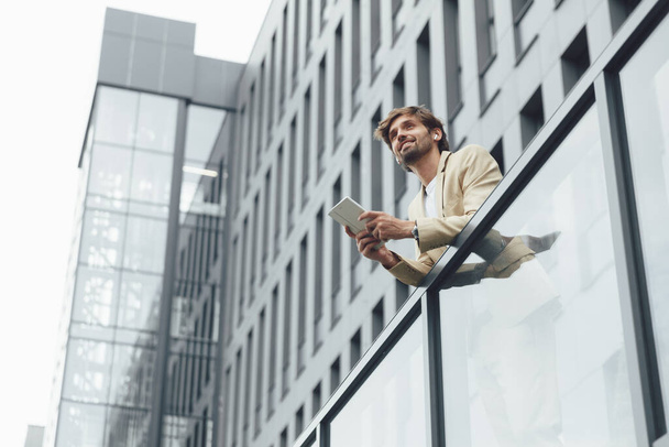 Portret van een bebaarde jonge zakenman met een digitale tablet terwijl hij op het balkon van zijn eigen kantoor staat. Begrip "mens", succes en technologie. - Foto, afbeelding