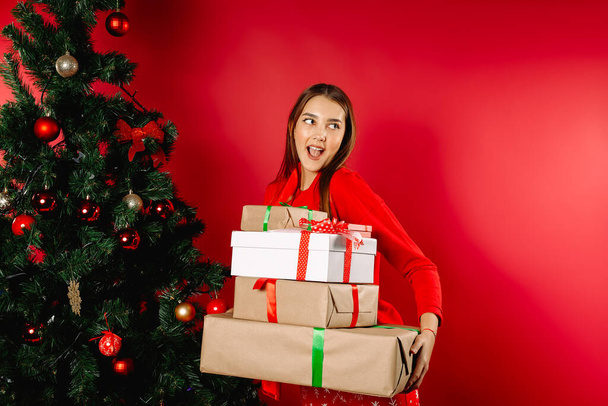 Surprised girl in pajamas holding a stack of gifts at home by the tree. The concept of New Year's gift, winter shopping. High quality photo - Fotografie, Obrázek