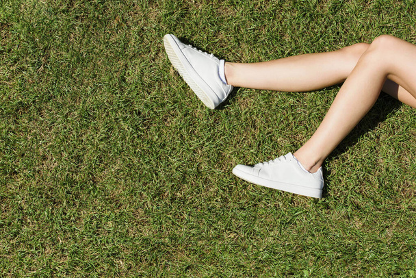 Female legs in white sneakers lie on the grass in a park. - Фото, зображення