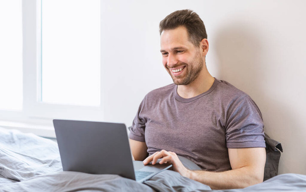 Χαρούμενος τύπος που χρησιμοποιεί το lap-top που εργάζονται σε απευθείας σύνδεση από απόσταση κάθεται στην κρεβατοκάμαρα - Φωτογραφία, εικόνα