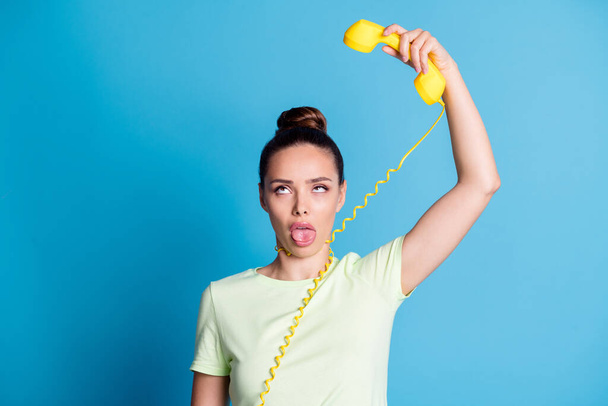 Φωτογραφία πορτρέτο της νεαρής grimacing κορίτσι δείχνει γλώσσα κρατώντας ρετρό τηλέφωνο με σύρμα γύρω από το λαιμό απομονώνονται σε έντονο μπλε χρώμα φόντο - Φωτογραφία, εικόνα