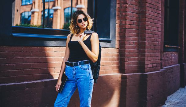 Stylish young woman in casual outfit and sunglasses with jacket on shoulder and notepad in hand standing on street near wall of modern building and looking away - Foto, Bild