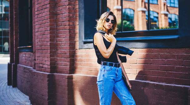 Confident female in summer outfit and sunglasses standing near red brick wall and mirror window with jacket on shoulder and folder in hands while walking on city street and looking at camera - Photo, image