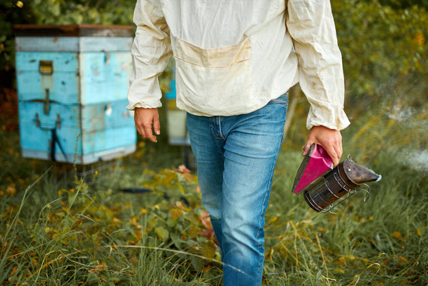 caucasian farmer tending his bee hives with bee smoker on his private farm with homemade bee boxes - Фото, изображение