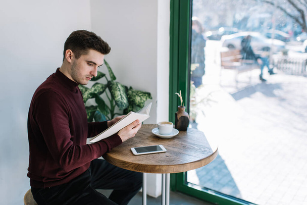 Intelligent male student reading journalistic publication while learning in cafe interior, smart man in casual wear spending time for studying and planning organisation in education textbook - Photo, image