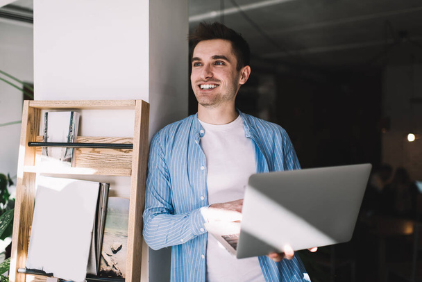 Cheerful Caucasian male copywriter smiling while waiting for laptop updating in coworking space, happy hipster guy feeling good during leisure for online shopping via modern netbook computer - 写真・画像