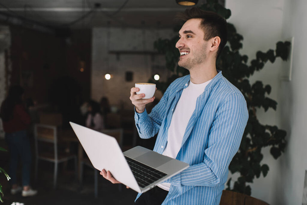 Side view of cheerful male in casual wear smiling and holding laptop and drinking cup of hot coffee in office - Fotografie, Obrázek