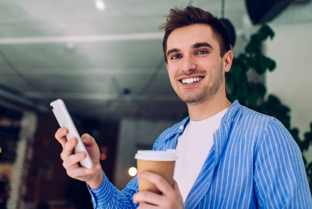 Half length portrait of cheerful male blogger with coffee to go and smartphone in hands smiling at camera, Caucasian millennial generation Z with takeaway cup enjoying networking via cellular app - Foto, immagini