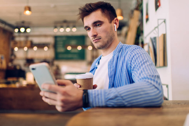 Back view of serious man with dark hair in casual clothes holding coffee and browsing smartphone while messaging with friends in cafeteria - Photo, Image