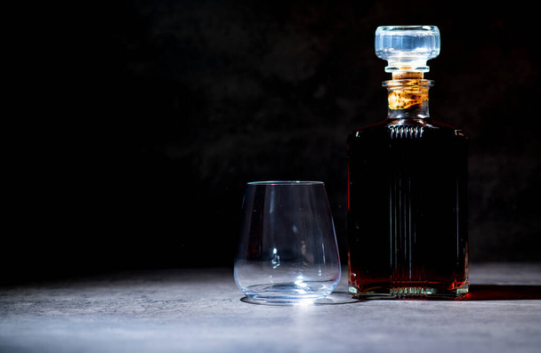 láhev whisky ve čtvercovém tvaru v paprscích světla vedle prázdné sklenice na tmavě šedém cementovém pozadí - Fotografie, Obrázek