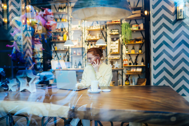 Красивая молодая женщина в повседневной одежде и в очках сидит одна за длинным деревянным столом с кружкой чая, разговаривая по телефону с ноутбуком в современном кафе Барселоны, глядя в сторону  - Фото, изображение