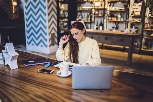 Стурбована прекрасною жінкою в окулярах, яка читає документи і банкноти, сидячи за столом з ноутбуком і чашкою в кафе в Барселоні - Фото, зображення