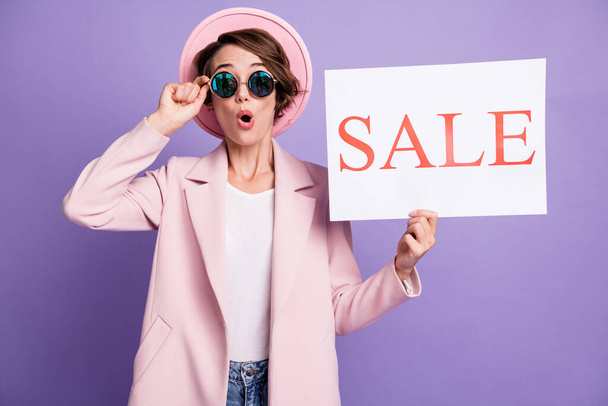 Photo of amazed pretty young lady holding in hand sale promo wear coat cap glasses isolated on pastel purple background - Foto, Bild