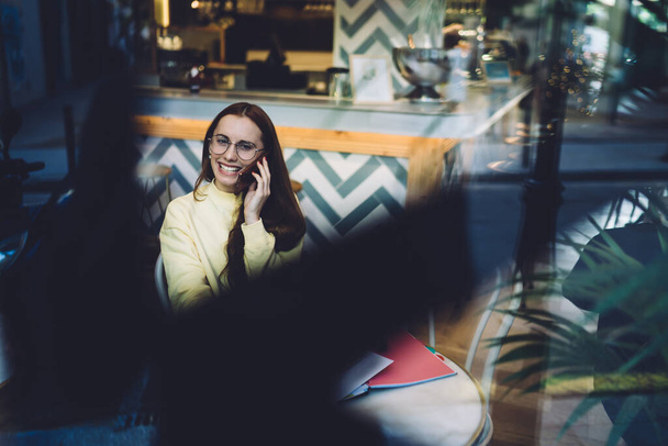 Young attractive female in trendy glasses and bright yellow sweatshirt talking on phone, smiling and looking at camera, while sitting in cozy cafe - Foto, Imagem