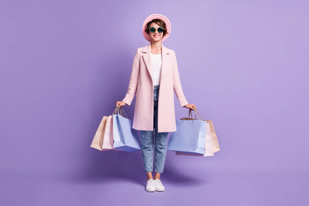 Full size photo of cheerful positive young woman stand hold purchases wear glasses pink coat jeans footwear isolated on purple background - Foto, Imagen